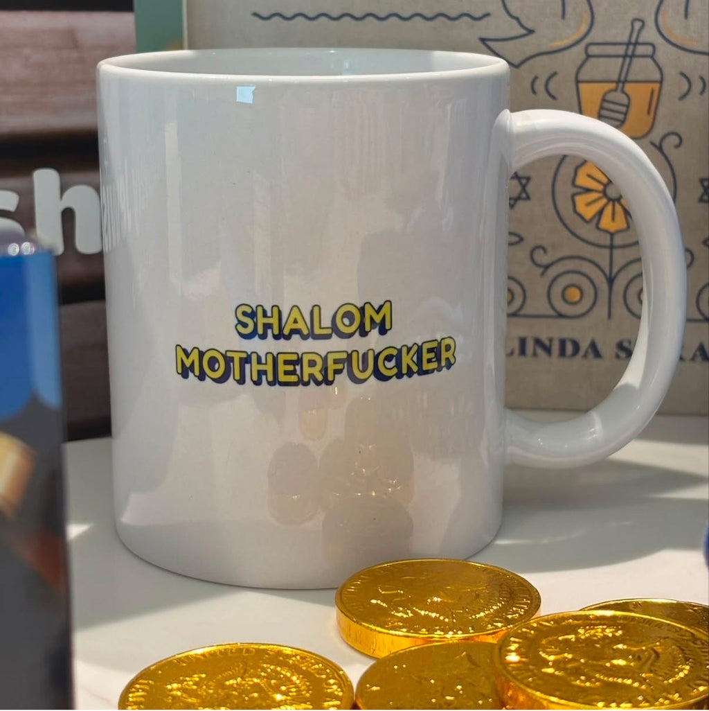 A white ceramic mug with the text 'SHALOM MOTHERFUCKER' printed on the side in yellow and blue, shown on a tabletop with gold chocolate coins in the foreground.