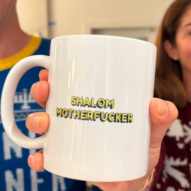 A white ceramic mug with the text 'SHALOM MOTHERFUCKER' printed in yellow with a black outline, held by a person in an indoor setting.