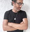 Man wearing a black t-shirt and glasses with a necklace, standing against a light textured wall.