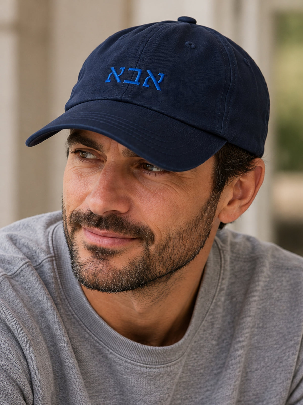 Man wearing a navy Abba (אבא) dad hat with bright blue Hebrew embroidery and a gray sweatshirt.