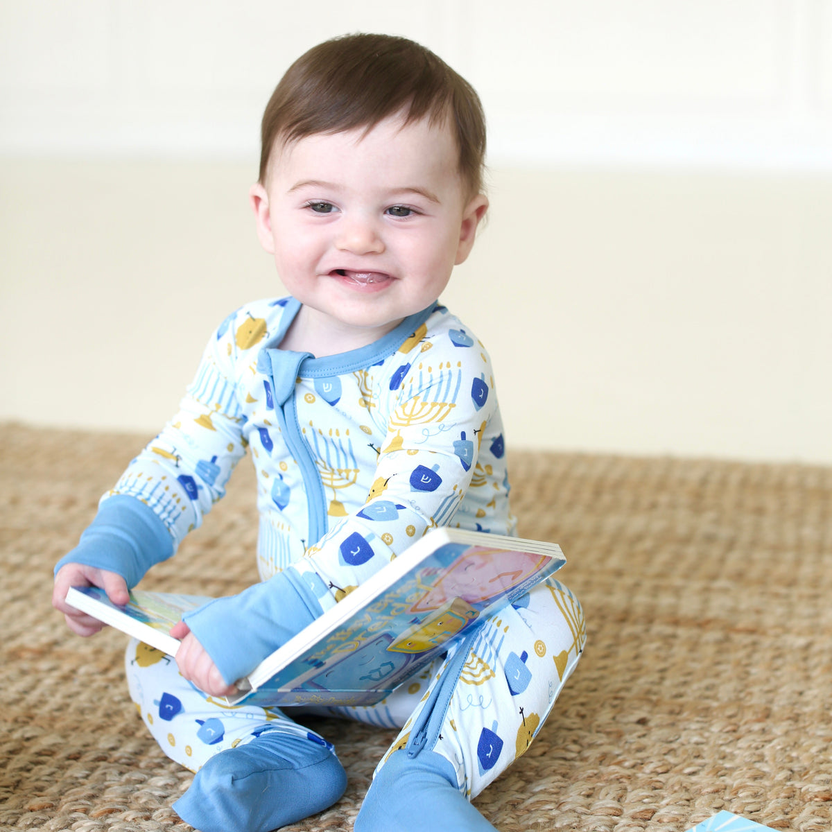 Smiling baby wearing the Latkes & Light Hanukkah Zippy while holding a book, with convertible feet folded closed.