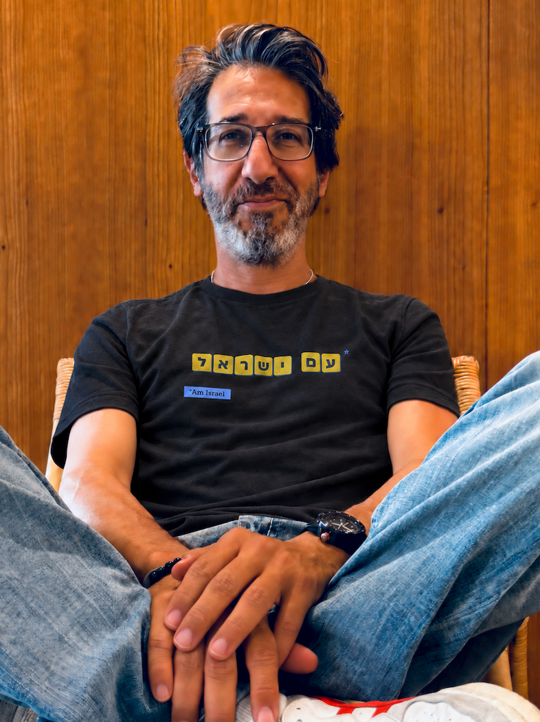 Eitan Chitayat wearing a black Am Yisrael T-shirt with yellow Hebrew lettering, seated against a wood-paneled wall.