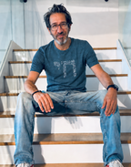 Eitan Chitayat seated on a staircase wearing a heather blue Chai T-shirt featuring the Hebrew word חי in a white dotted design, paired with light-wash jeans.