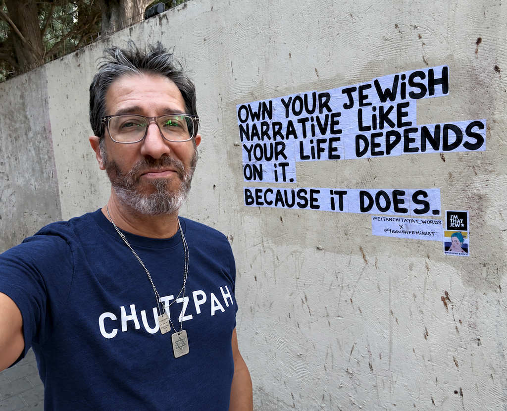 Eitan Chitayat wearing a navy Chutzpah T-shirt with white lettering, standing beside a wall displaying the message “Own your Jewish narrative like your life depends on it. Because it does.”