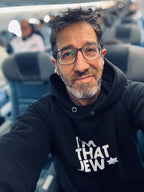 Eitan Chitayat wearing a black I'm That Jew hoodie with bold white lettering and a Star of David, seated on an airplane.
