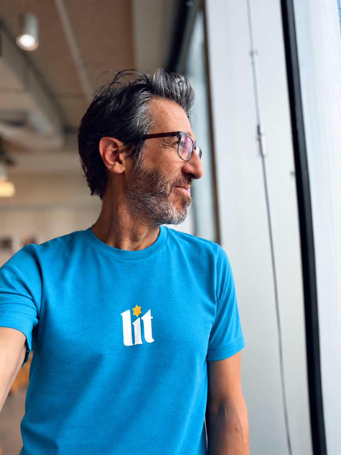 Eitan Chitayat wearing a turquoise Lit T-shirt with a small multicolored chest graphic, standing beside a window.