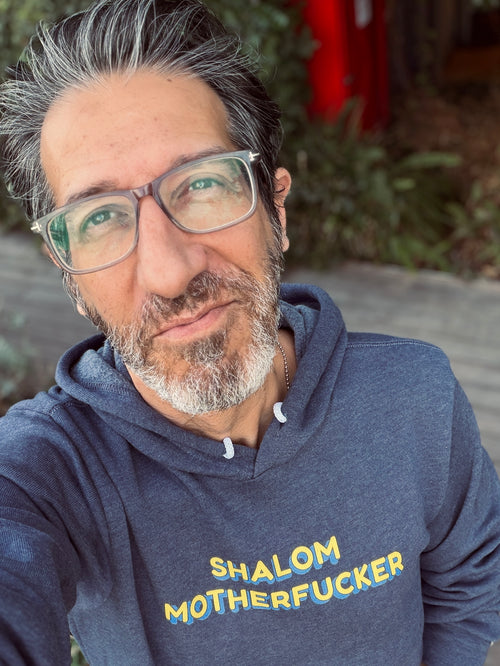 Eitan Chitayat wearing a heather navy Shalom Motherfucker hoodie with yellow and blue lettering, photographed outdoors in a close-up selfie.