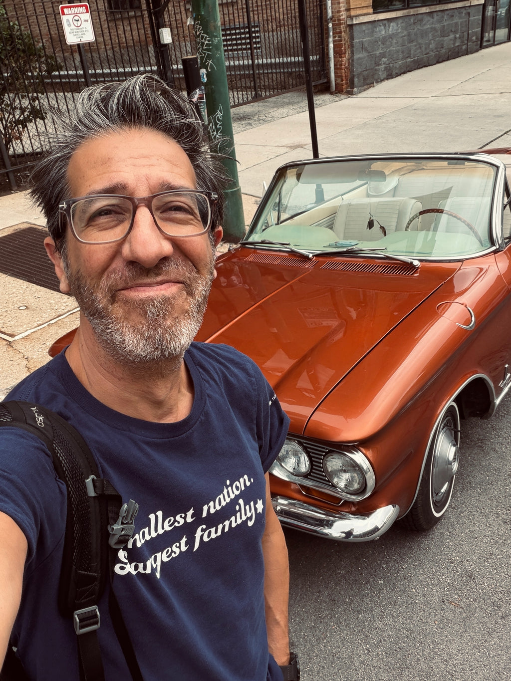 Eitan Chitayat wearing a navy Smallest Nation, Largest Family T-shirt with white lettering, taking a selfie beside a vintage copper-colored convertible.