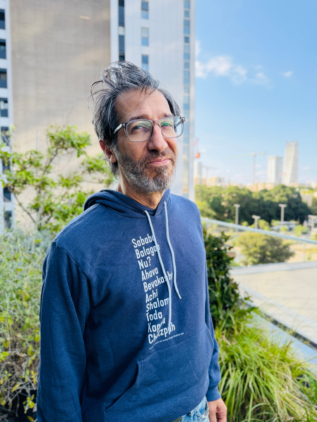Eitan Chitayat wearing a heather blue The Ten Hebrew Words hoodie with white lettering listing Hebrew expressions including Sababa, Balagan, Nu, Ahava, B'vakasha, Achi, Shalom, Toda, Kapara, and Chutzpah, photographed outdoors.