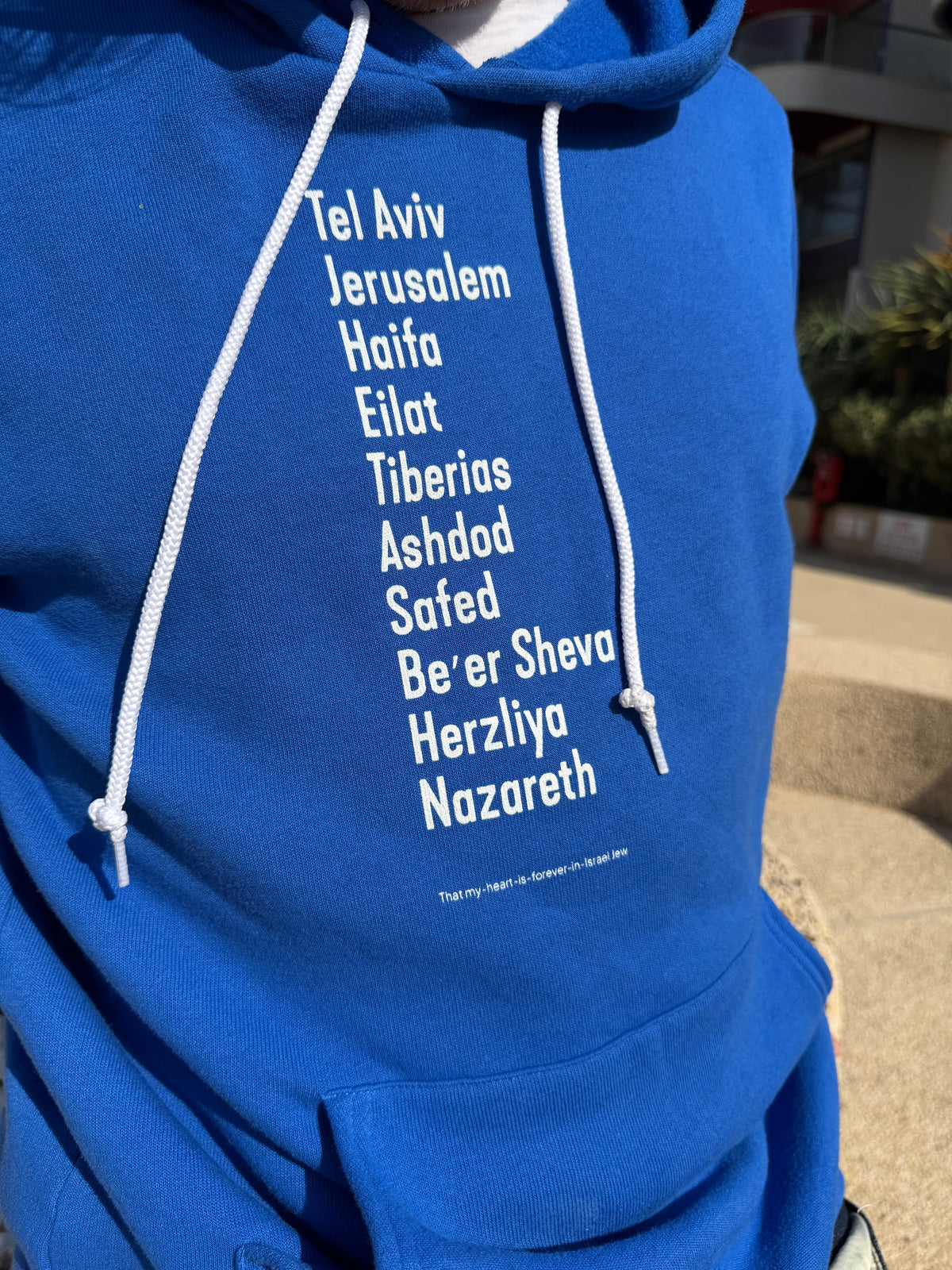 Eitan Chitayat wearing a royal blue The Ten Israeli Cities hoodie with white lettering listing Tel Aviv, Jerusalem, Haifa, Eilat, Tiberias, Ashdod, Safed, Be'er Sheva, Herzliya, and Nazareth.