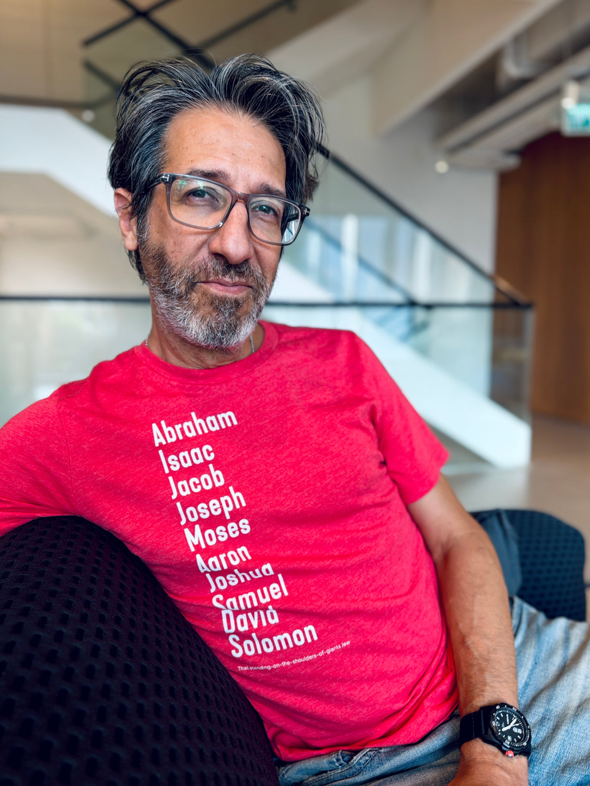 Eitan Chitayat wearing a heather red The Ten Patriarchs T-shirt with white lettering listing Abraham, Isaac, Jacob, Joseph, Moses, Aaron, Joshua, Samuel, David, and Solomon, seated indoors.