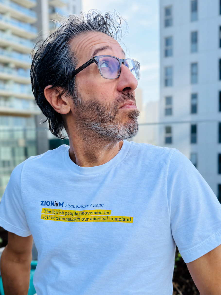 Eitan Chitayat wearing a white ZIONisM Definition T-shirt with blue lettering and a yellow-highlighted definition, photographed outdoors against a city backdrop.