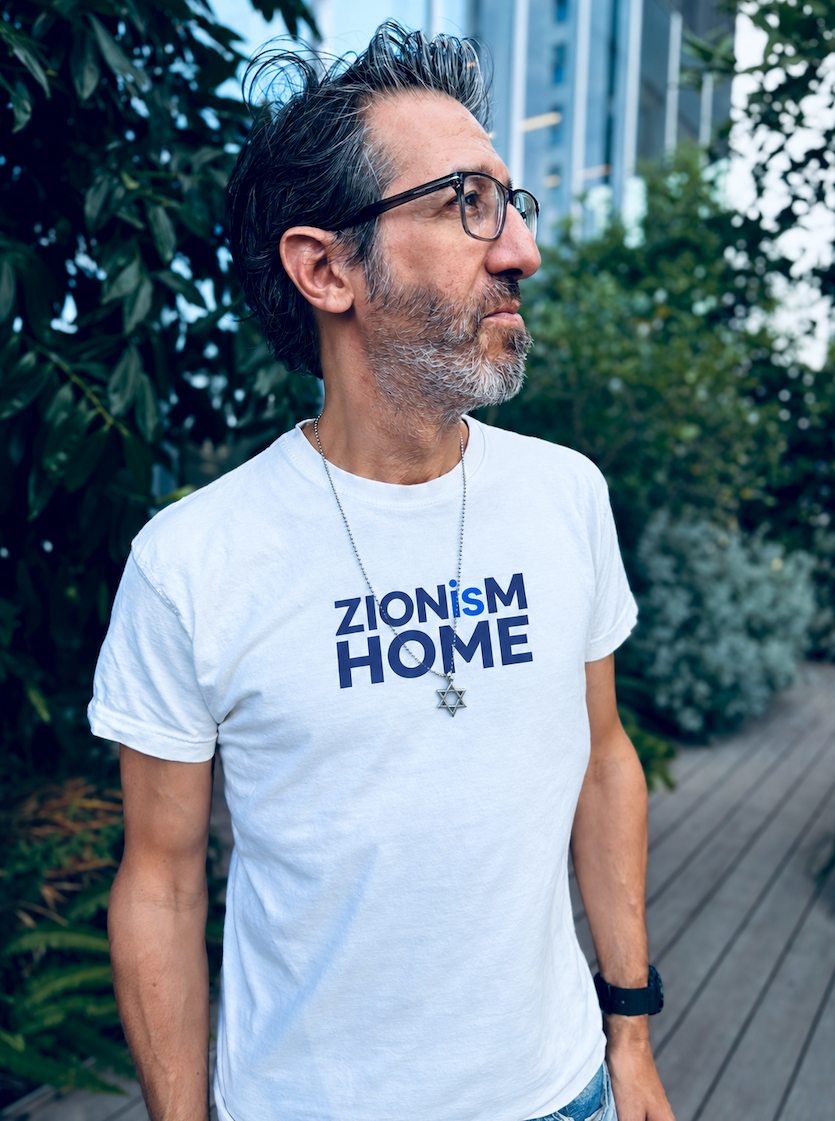 Eitan Chitayat wearing a white ZIONisM Home T-shirt with navy blue lettering, standing outdoors on a wooden walkway surrounded by greenery.