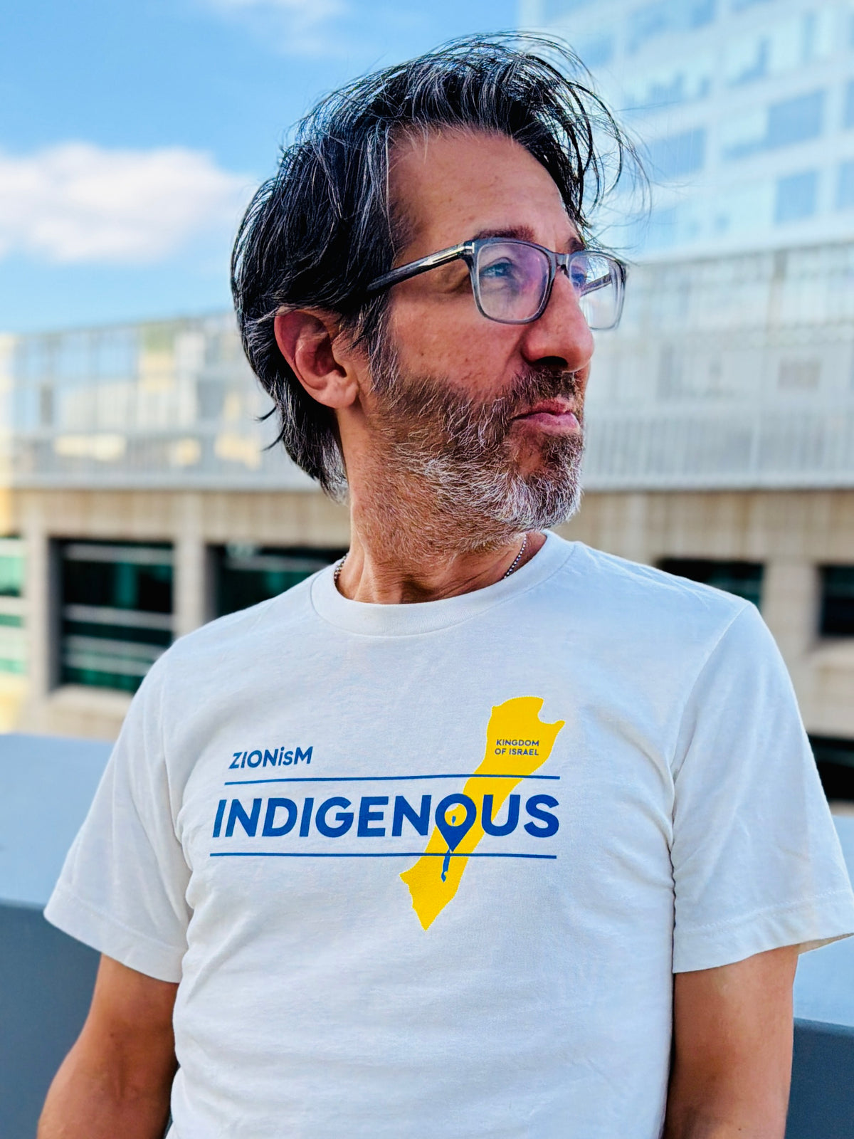 Eitan Chitayat wearing a vintage white ZIONisM Indigenous T-shirt featuring blue lettering and a yellow map of Israel, photographed outdoors against a modern building.