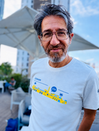 Eitan Chitayat wearing a white ZIONisM Shalom T-shirt featuring blue and yellow Israeli-inspired artwork and the Hebrew word שלום, photographed outdoors.