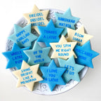 Blue, aqua, and cream Star of David marzipan conversation tiles featuring playful Hanukkah sayings arranged on a serving plate.