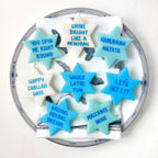 Star of David marzipan conversation tiles in blue, aqua, and cream with playful Hanukkah sayings arranged on a serving plate.