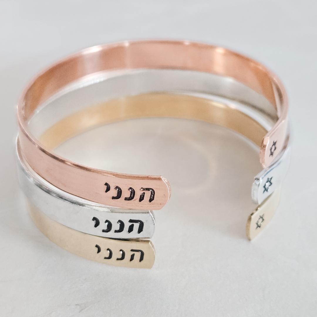 Three Hineni Hebrew cuff bracelets in rose gold, sterling silver, and gold finishes, hand-stamped with הנני and featuring small Star of David details.