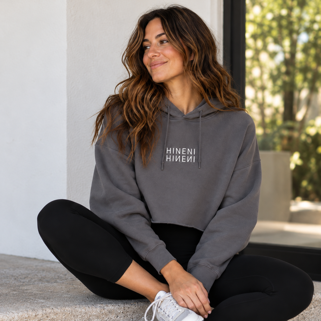 Woman seated cross-legged on stone steps wearing a charcoal gray HINENI reflection cropped hoodie with white mirrored lettering, black leggings, and white sneakers.