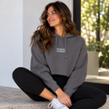 Woman seated cross-legged on stone steps wearing a charcoal gray HINENI reflection cropped hoodie with white mirrored lettering, black leggings, and white sneakers.