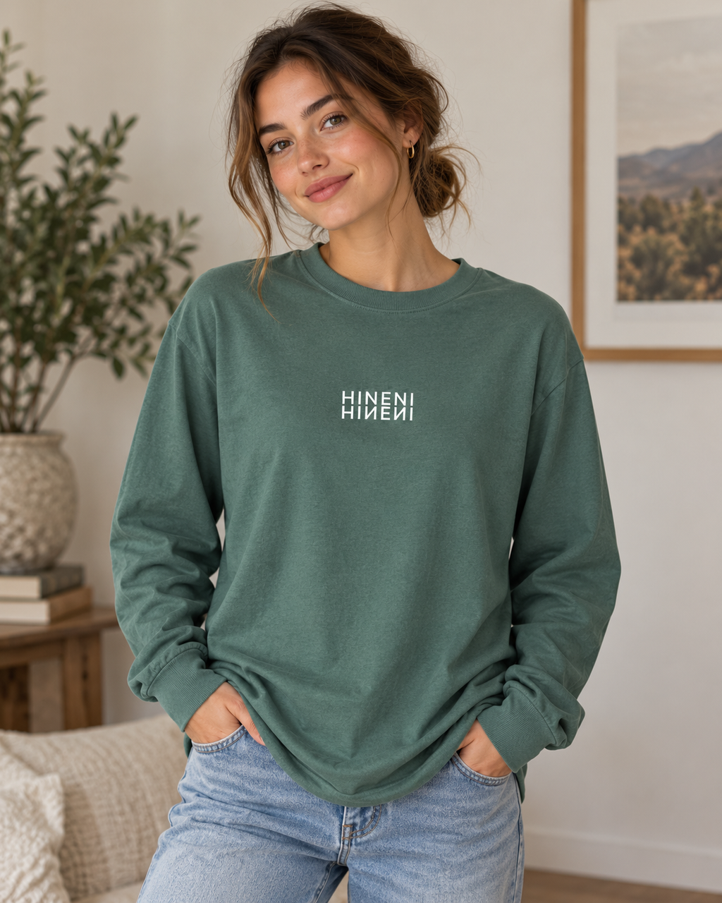 Woman wearing the Hineni Reflection heavyweight garment-dyed long-sleeve tee in Washed Pine, featuring white mirrored HINENI lettering across the chest, styled with light-wash jeans.