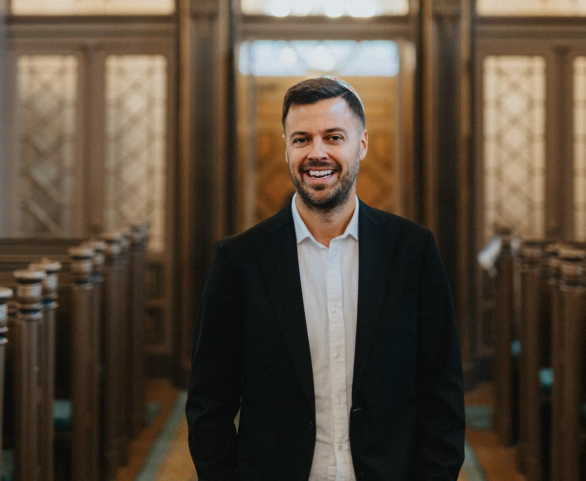 A smiling man with a beard and a white kippah stands in a wooden synagogue or chapel, wearing a black blazer over a white button-down shirt.