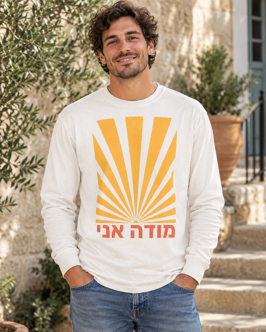 Man wearing the Modeh Ani Sunrise long-sleeve T-shirt in Vintage White, featuring a golden sunrise graphic and coral Hebrew “Modeh Ani” text on the front.