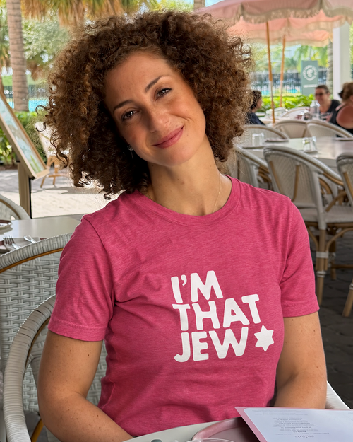 Monica Czukerberg wearing a heather raspberry I'm That Jew T-shirt with bold white lettering and a Star of David, seated at an outdoor café.
