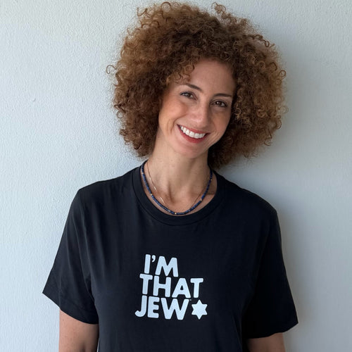 A smiling woman with curly brown hair wearing a black t-shirt with the text 'I'M THAT JEW' printed in white next to a white star, standing against a light gray wall.