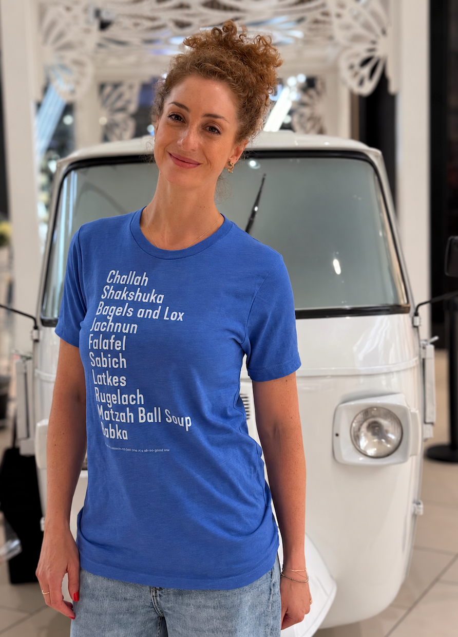 Monica Czukerberg wearing a blue The Ten Jewish Foods T-shirt featuring a list of Jewish dishes in white lettering, standing in front of a vintage white vehicle.
