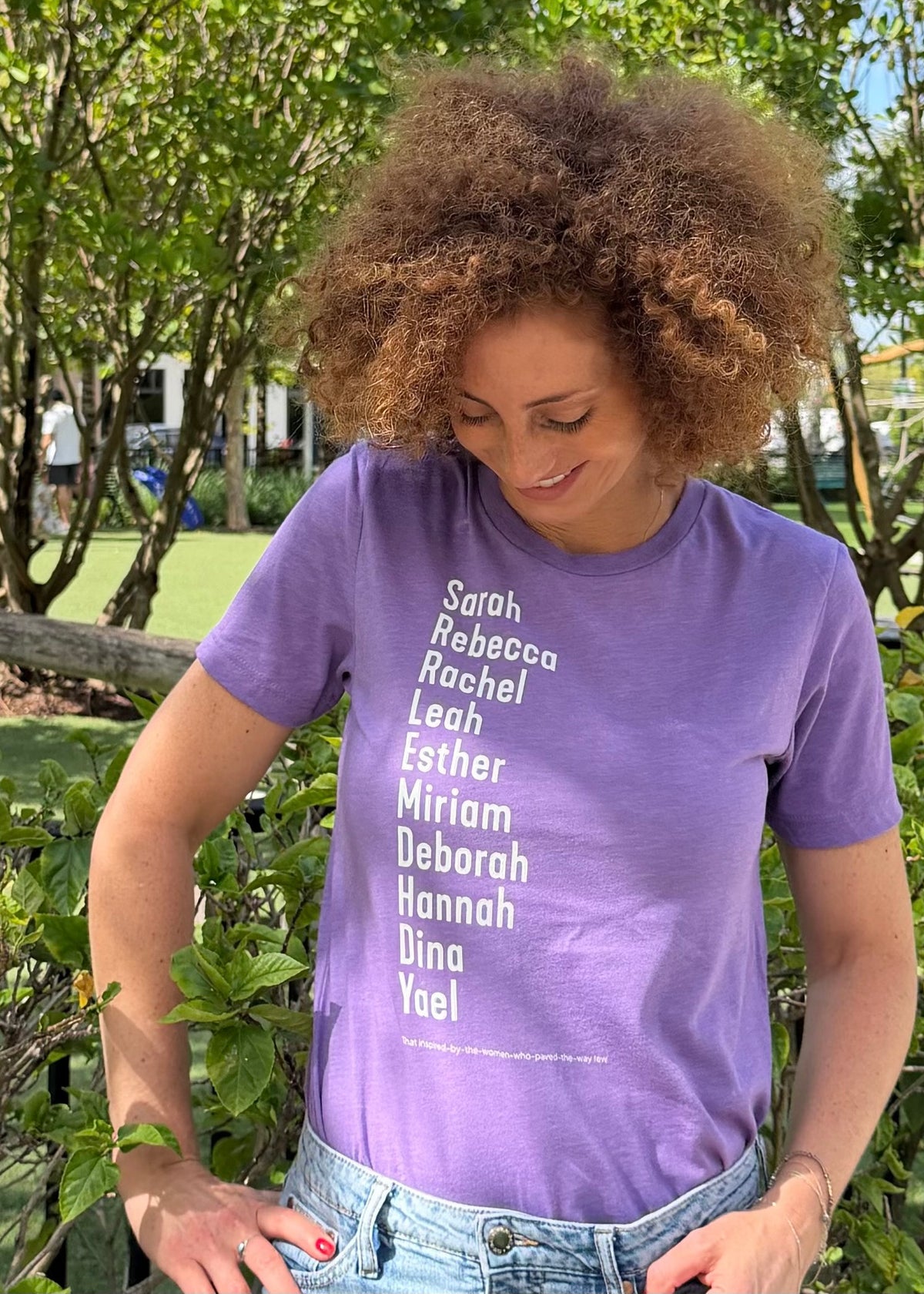 Monica Czukerberg wearing a purple The Ten Matriarchs T-shirt with white lettering listing Sarah, Rebecca, Rachel, Leah, Esther, Miriam, Deborah, Hannah, Dina, and Yael, standing outdoors among greenery.