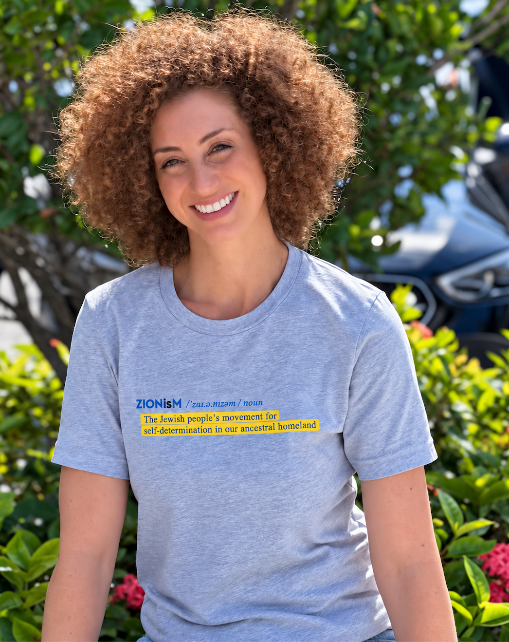 Monica Czukerberg wearing a heather gray ZIONisM Definition T-shirt with blue lettering and a yellow-highlighted definition, photographed outdoors.