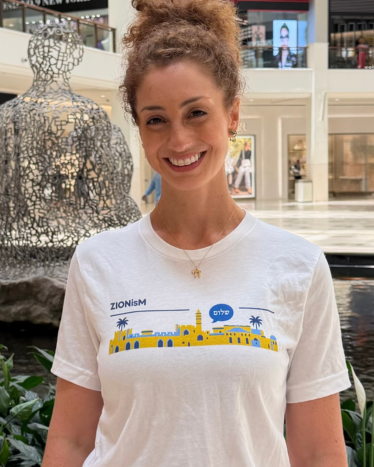 Monica Czukerberg wearing a white ZIONisM Shalom T-shirt featuring a blue and yellow Jerusalem skyline and the Hebrew word שלום, photographed indoors.