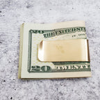 Yellow brass Nice Jewish Boy money clip holding a folded stack of twenty-dollar bills, showing how the clip secures cash.