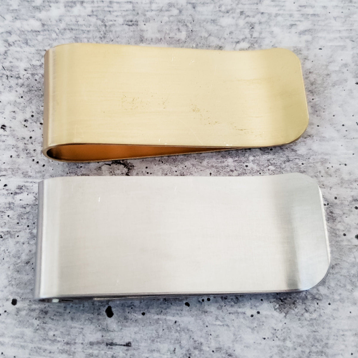 Back view of two handmade money clips in yellow brass and silver finishes, showing their smooth metal surfaces and folded edges.