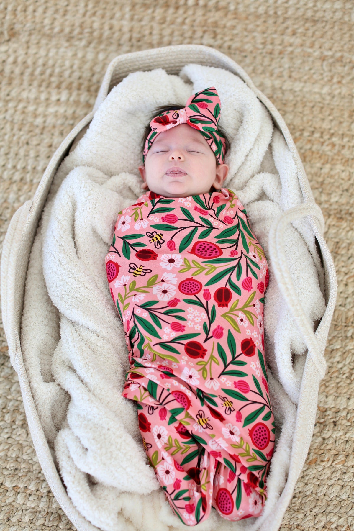 Newborn in a basket wrapped in the pink Petals & Pomegranates swaddle with matching bow