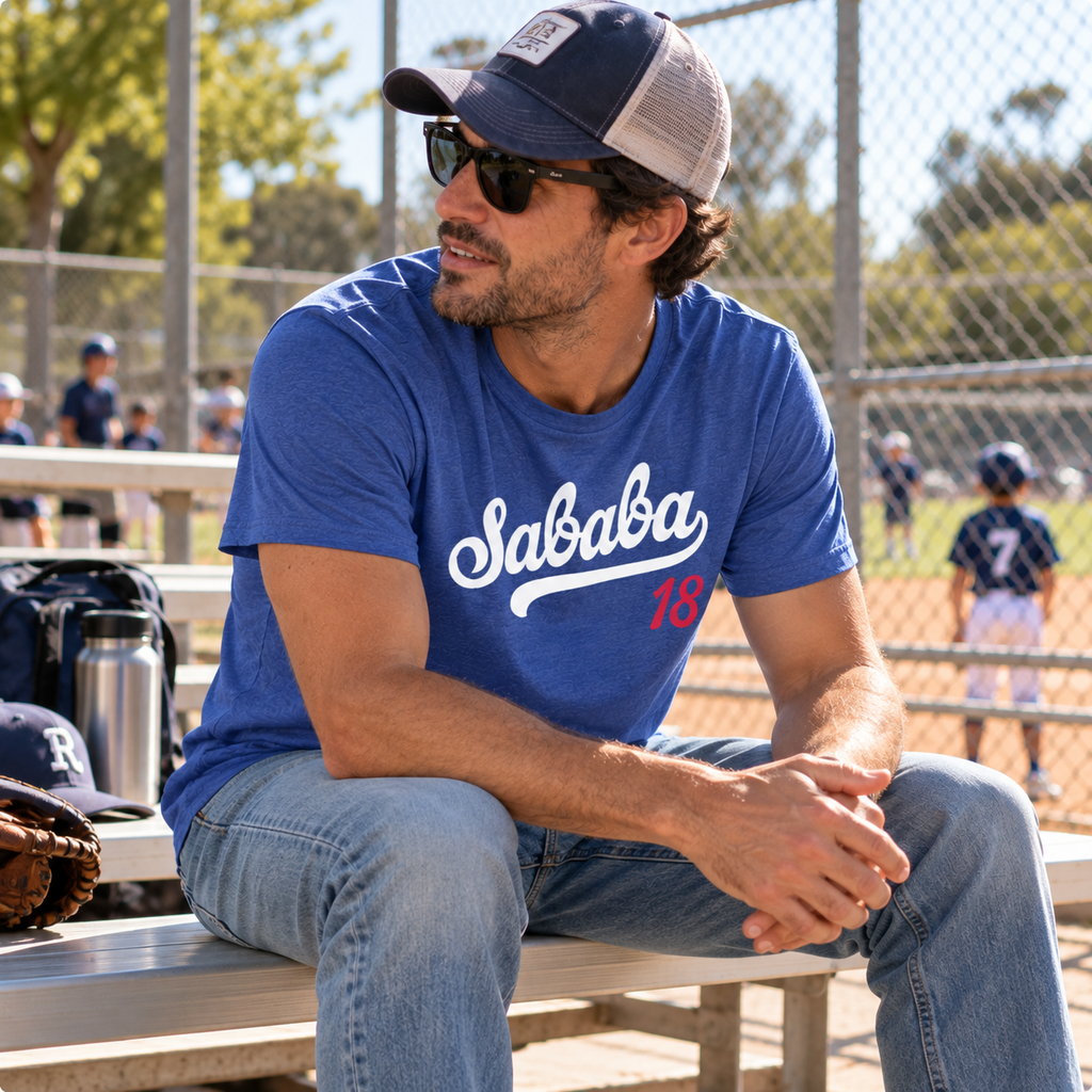 Man wearing a blue Sababa 18 T-shirt while sitting on bleachers at a baseball field.