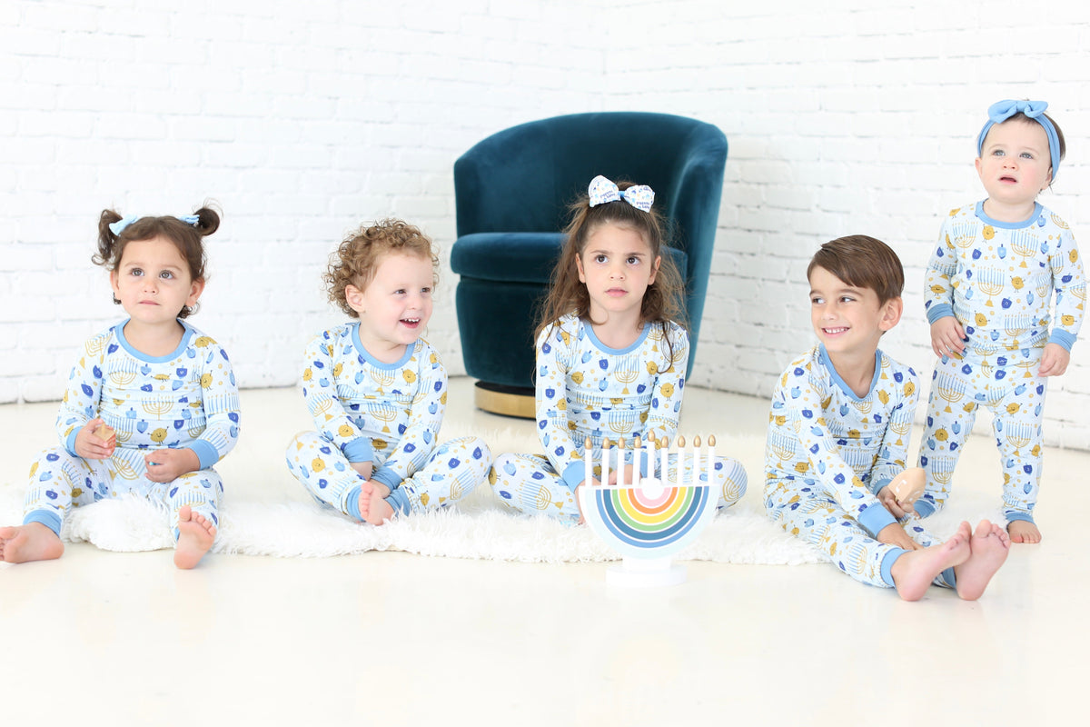 Five children wearing Shiloh & Kate Latkes & Light Hanukkah pajamas gathered around a rainbow menorah.