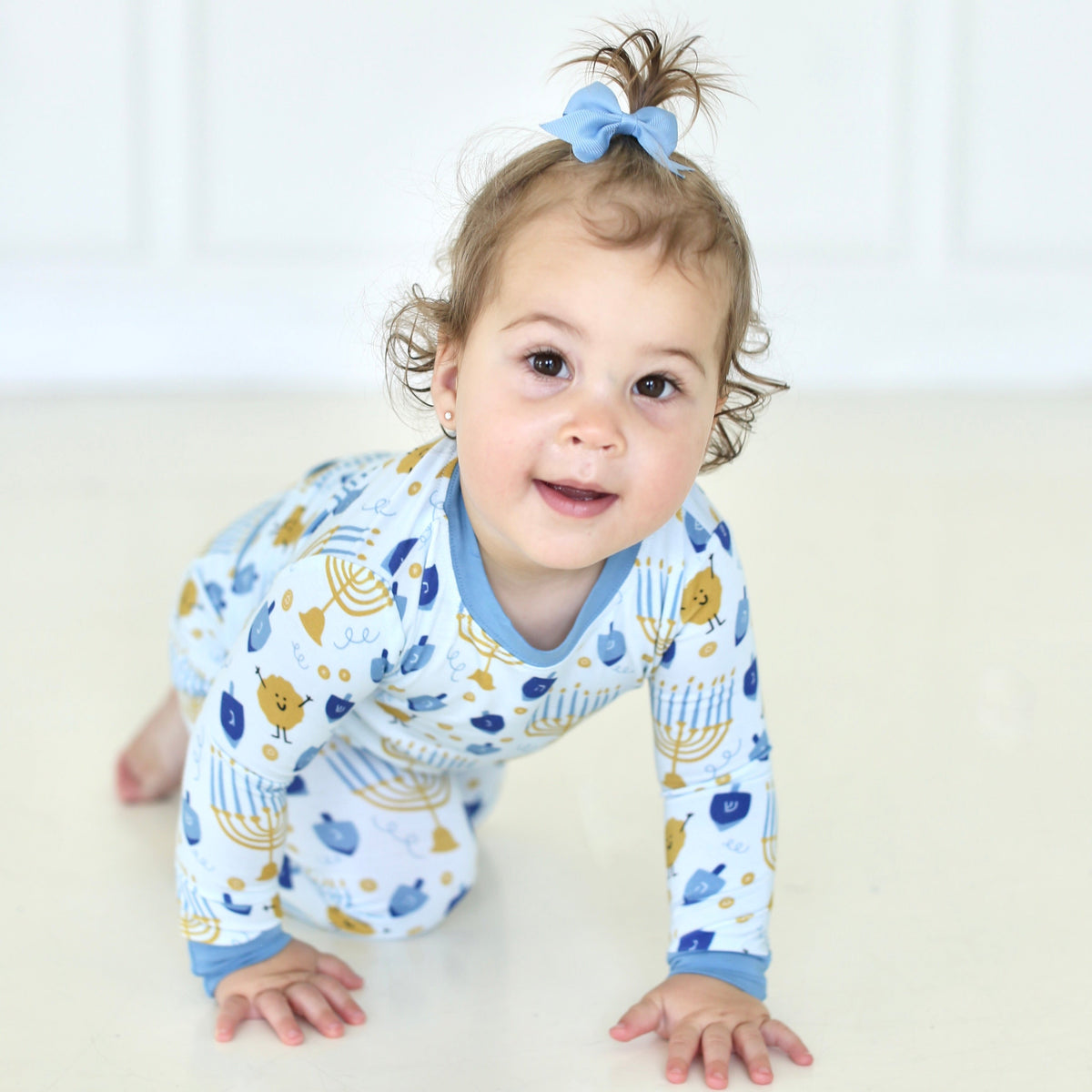 Toddler crawling in Shiloh & Kate Latkes & Light Hanukkah pajamas with blue trim and menorah, dreidel, and latke illustrations.
