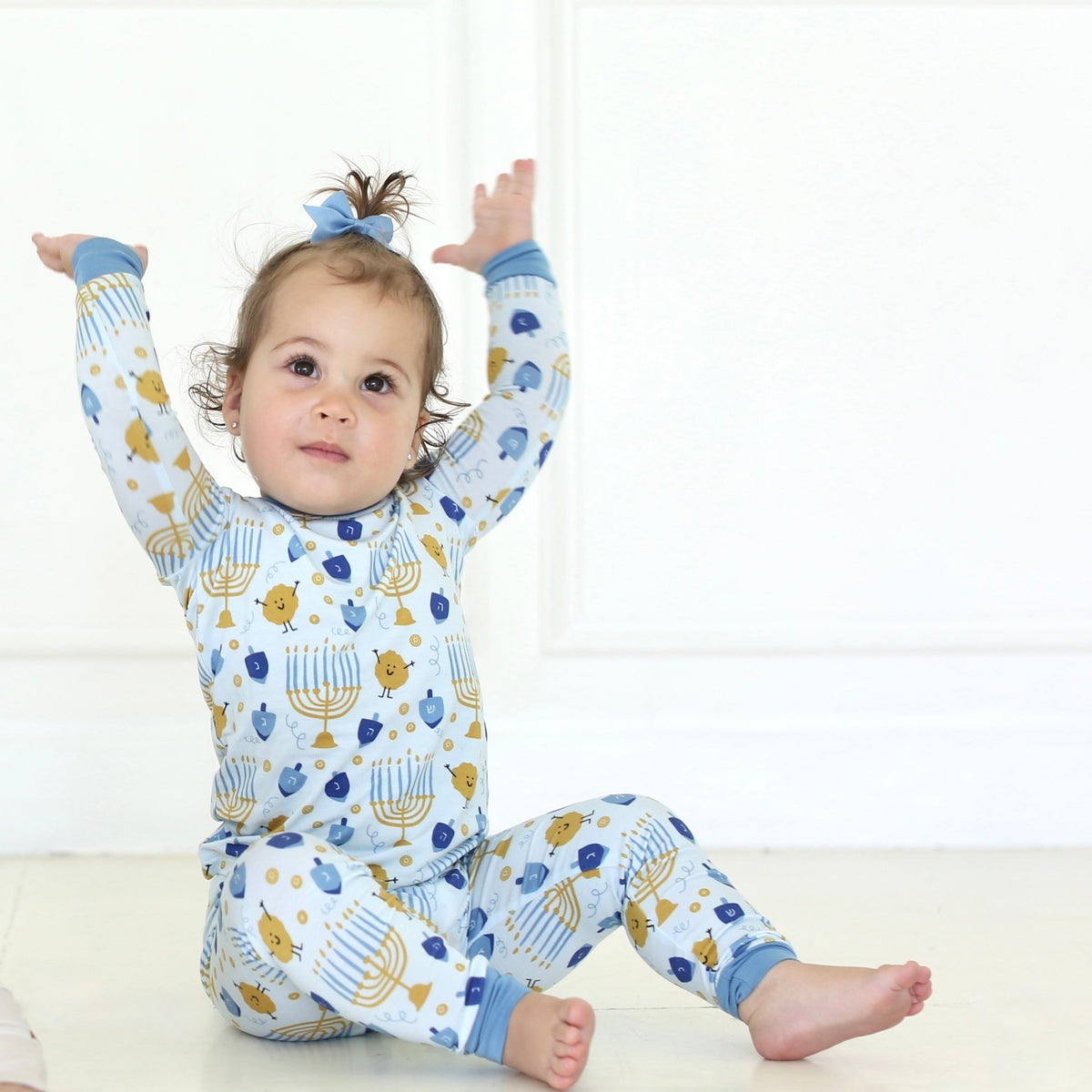 Toddler seated with arms raised, wearing Shiloh & Kate Latkes & Light two-piece Hanukkah pajamas with blue trim.