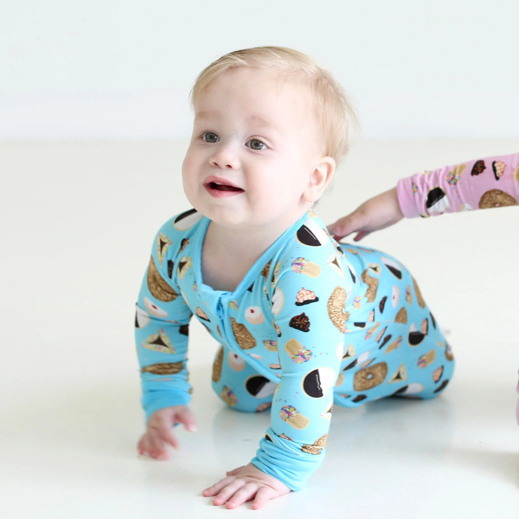 Baby crawling in Shiloh & Kate Blue Jewish Baked Goods zip-up pajamas, featuring bagels, challah, and assorted Jewish treats on a bright blue background.