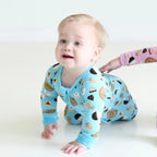 Baby crawling in Shiloh & Kate Blue Jewish Baked Goods zip-up pajamas, featuring bagels, challah, and assorted Jewish treats on a bright blue background.