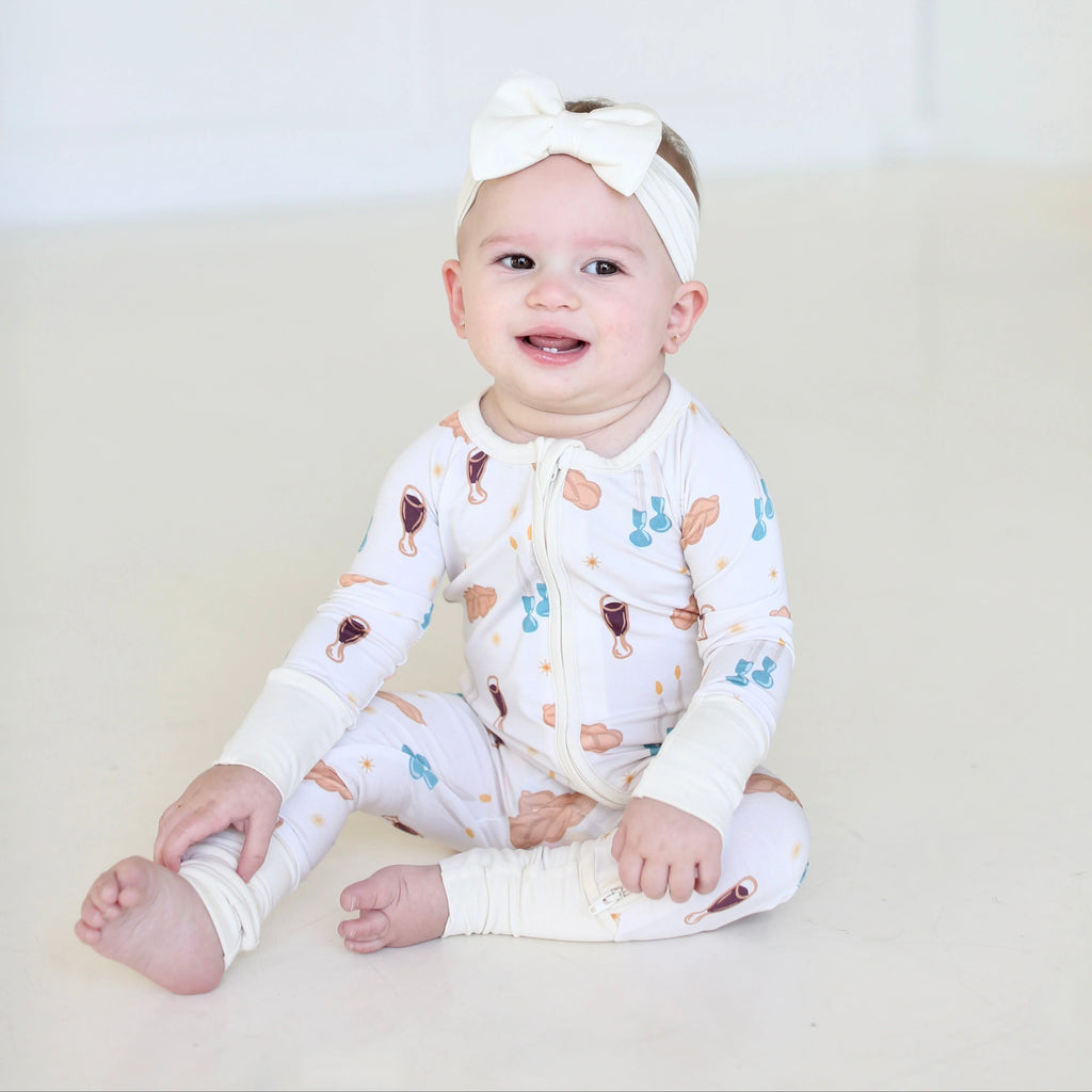 Smiling baby wearing Shiloh & Kate Cream Shabbat zip-up pajamas with challah, Kiddush cups, and Shabbat candles, paired with a white bow headband.