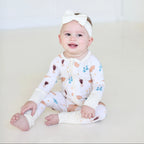 Smiling baby wearing Shiloh & Kate Cream Shabbat zip-up pajamas with challah, Kiddush cups, and Shabbat candles, paired with a white bow headband.