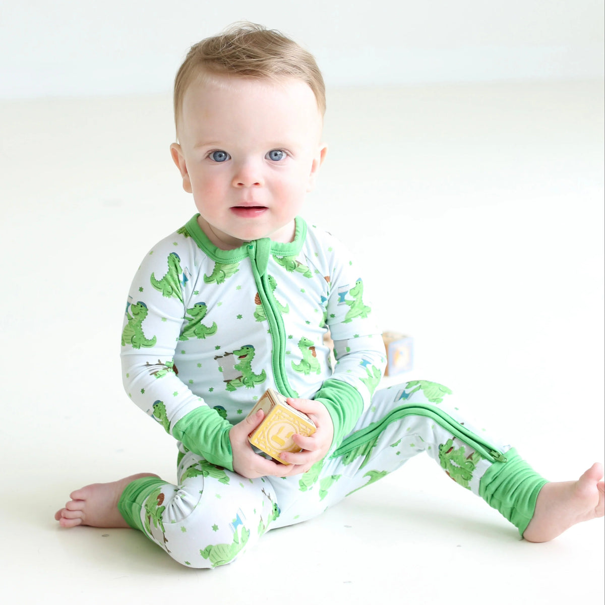 Baby wearing Shiloh & Kate Dinosaur Shabbat zip-up pajamas with playful green dinosaurs and matching green trim, sitting with a wooden toy block.