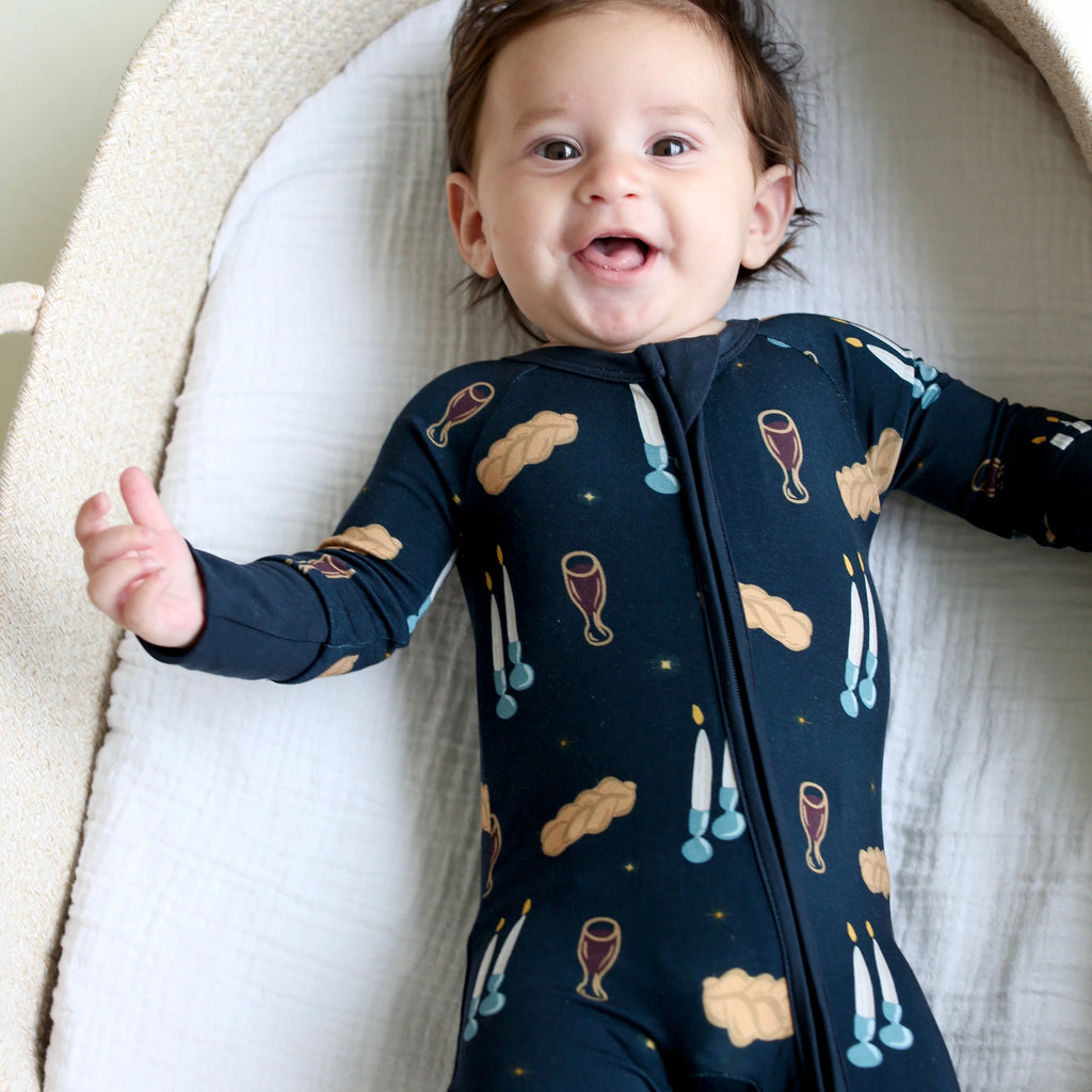 Smiling baby wearing Shiloh & Kate Navy Shabbat zip-up pajamas with challah, Kiddush cups, and Shabbat candles on a dark navy background.
