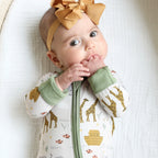 Baby wearing Shiloh & Kate Noah’s Ark zip-up pajamas featuring giraffes, Noah’s ark, and animals, with sage green trim and a mustard yellow bow headband.