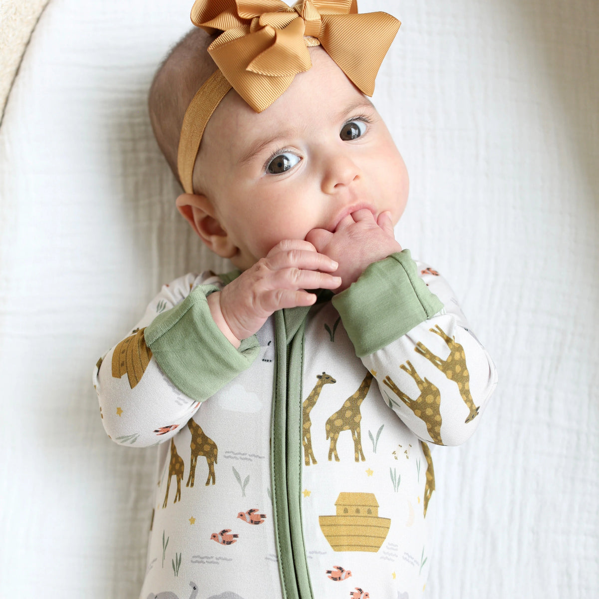 Baby wearing Shiloh & Kate Noah’s Ark zip-up pajamas featuring giraffes, Noah’s ark, and animals, with sage green trim and a mustard yellow bow headband.