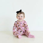 Smiling baby wearing Shiloh & Kate Pink Jewish Baked Goods zip-up pajamas featuring challah, bagels, and other Jewish treats, paired with a black bow headband.