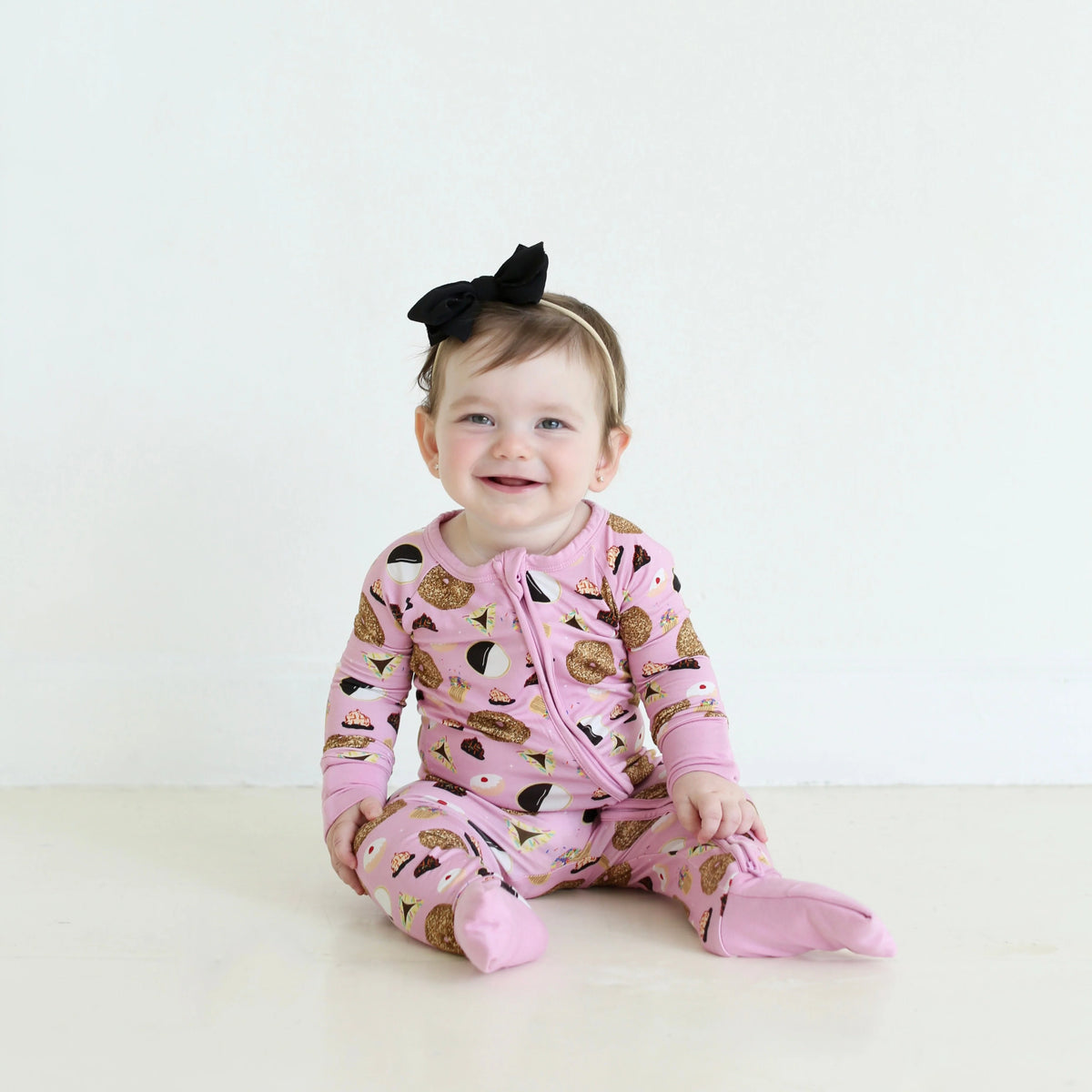 Smiling baby wearing Shiloh & Kate Pink Jewish Baked Goods zip-up pajamas featuring challah, bagels, and other Jewish treats, paired with a black bow headband.